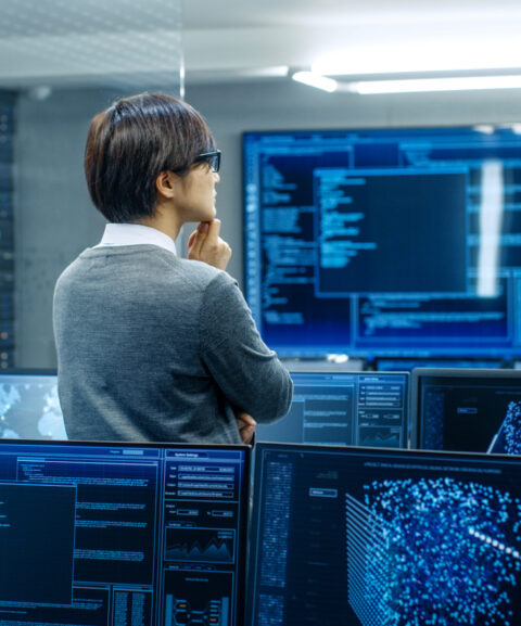 In the System Control Room Technical Operator Stands and Monitors Various Activities Showing on Multiple Displays with Graphics. Administrator Monitors Work of  Artificial Intelligence, Big Data Mining, Neural Network, Surveillance Project.